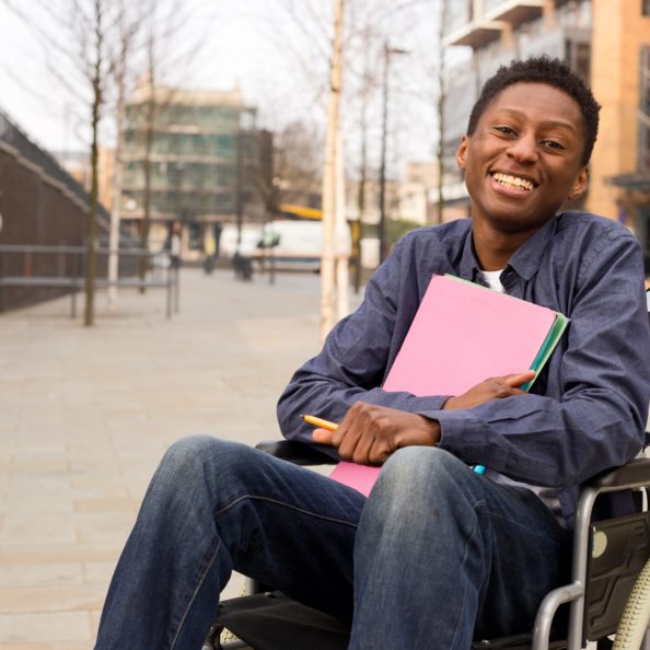 A teenager, holding hardcover books and pencil, positioned in front of building on a pedestrian walkway. He is using a folding-frame manual wheelchair.